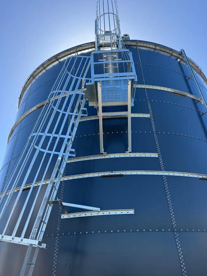 Low angle up the wall of a finished tank to the caged ladder and roof platform against a clear sky