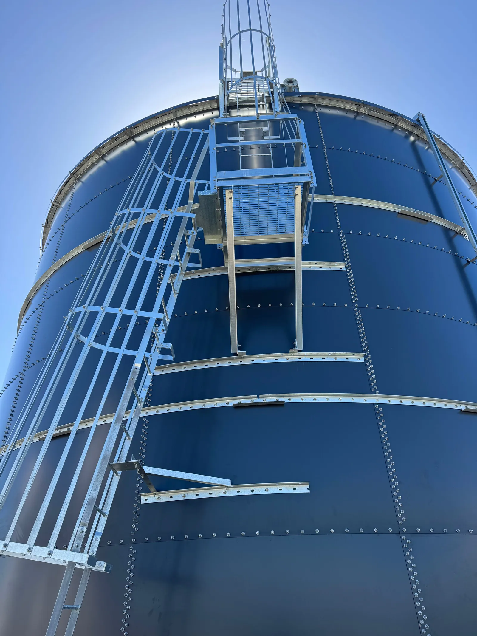 Low angle up the wall of a finished tank to the caged ladder and roof platform against a clear sky