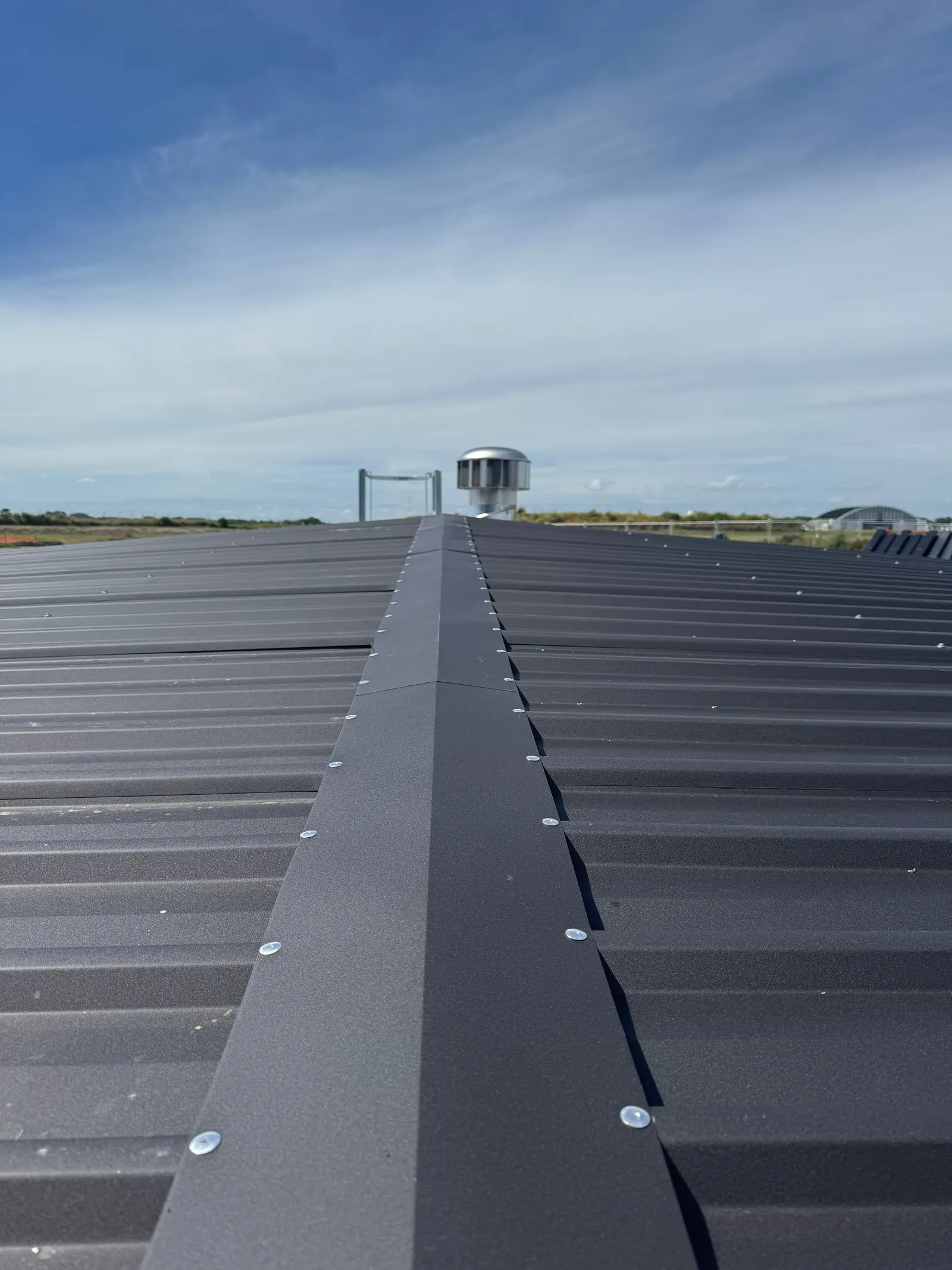 Along the ridge capping of a completed tank roof, turbine vent and roof hatch frame at the far end