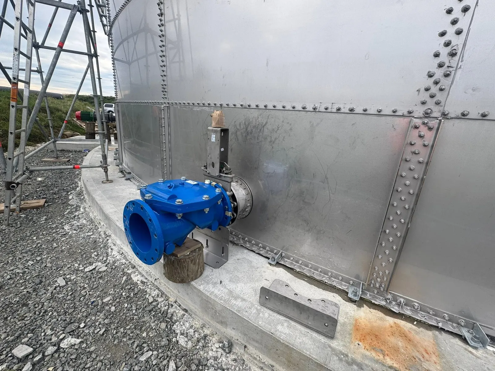 A blue cast iron outlet valve bolted to the wall of a galvanised tank on its concrete ring beam