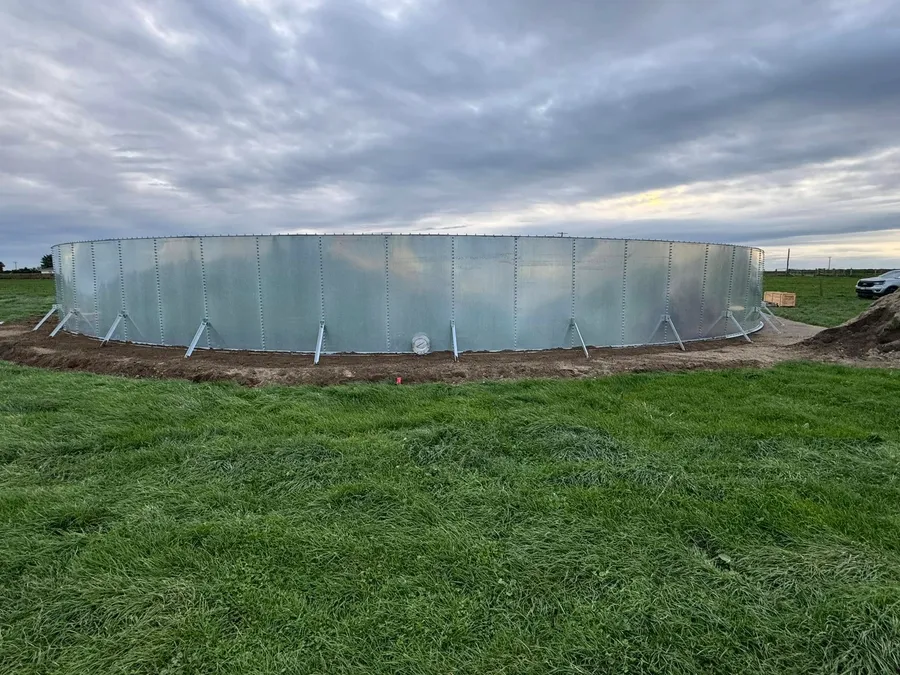 The completed galvanised bolted steel tank standing in a grass paddock under a heavy evening sky