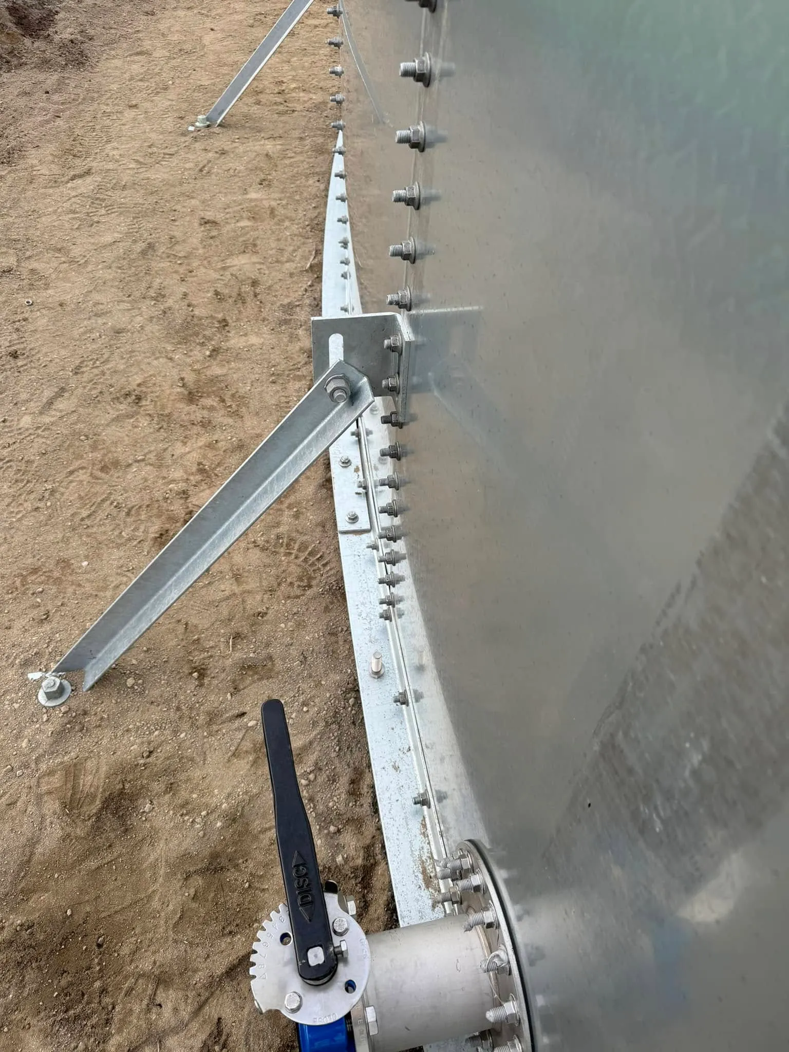 The base of a galvanised tank wall from above, anchor brackets and bracing set into the ground beside a butterfly valve