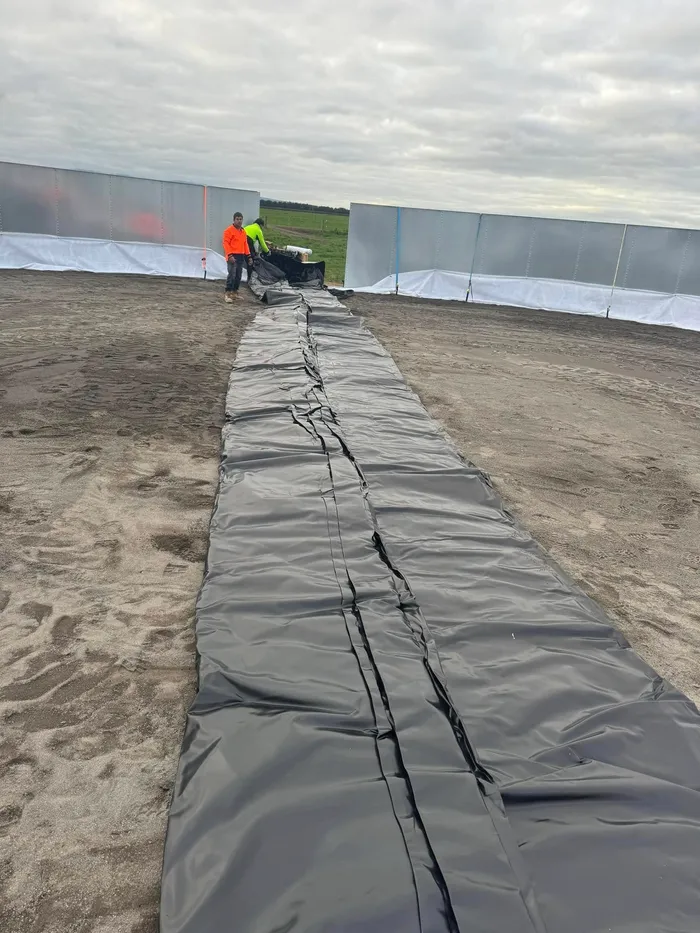 Black liner rolled out across the sand floor inside a part-erected tank shell, two crew unrolling it at the far wall