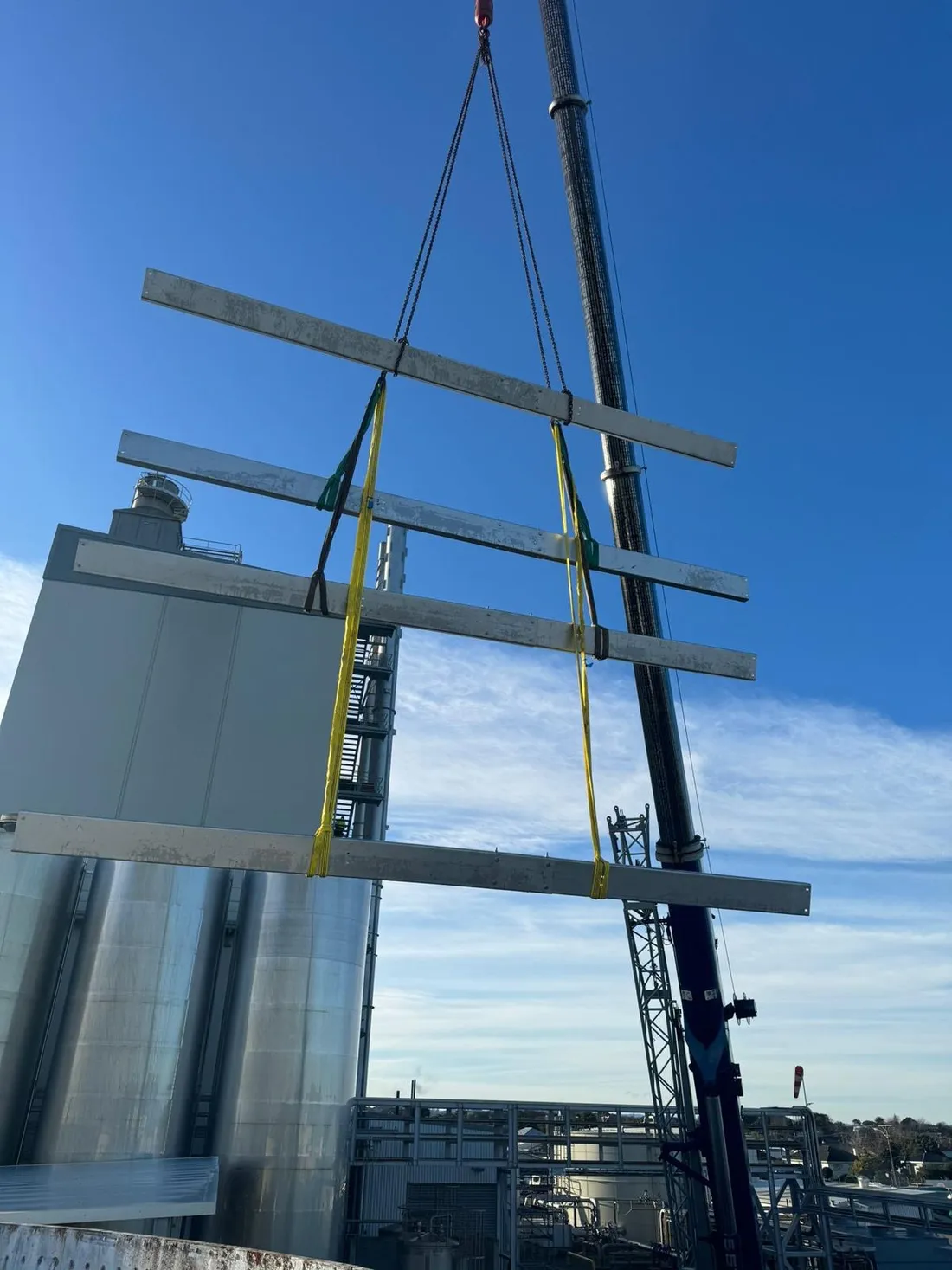 Steel roof beams hanging from a crane hook in clear sky above a tank site