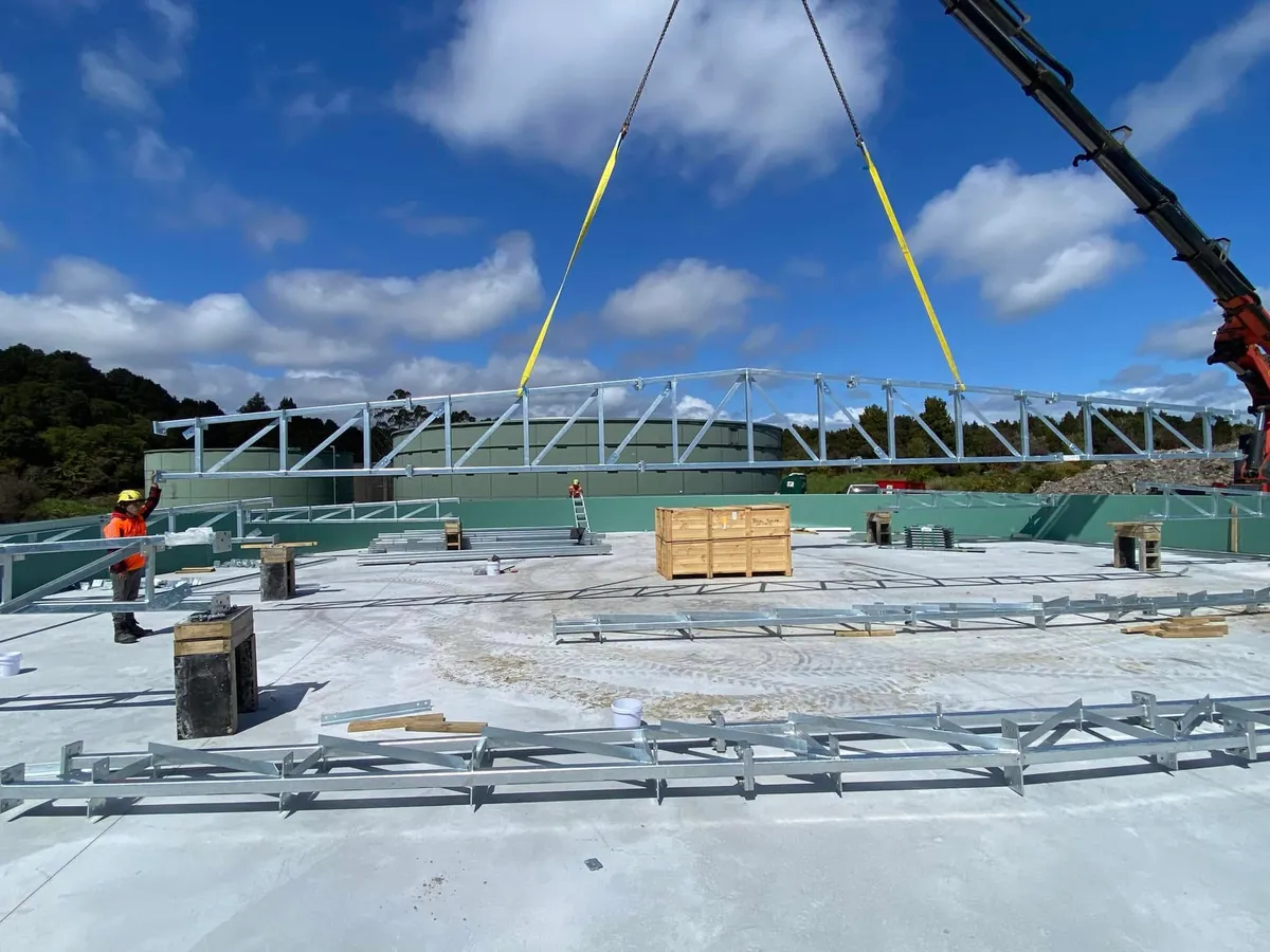 A crane lifting a galvanised roof truss over a tank under construction