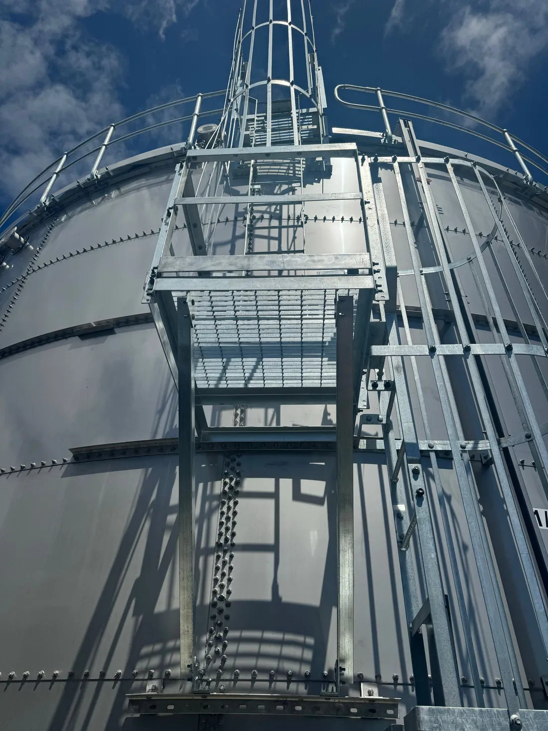 Low angle view up a galvanised access stair and caged ladder fixed to a white bolted steel tank