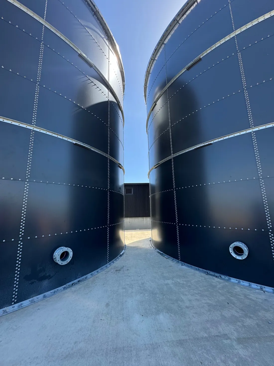 Two navy bolted steel tanks standing side by side on a concrete pad, photographed from the gap between them