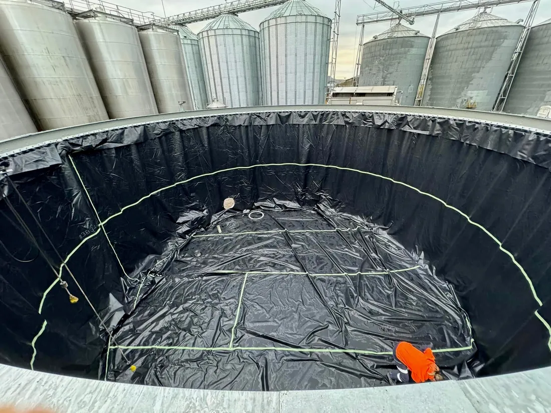 Crew member working on a black floor liner inside a large tank shell, grain silos standing behind