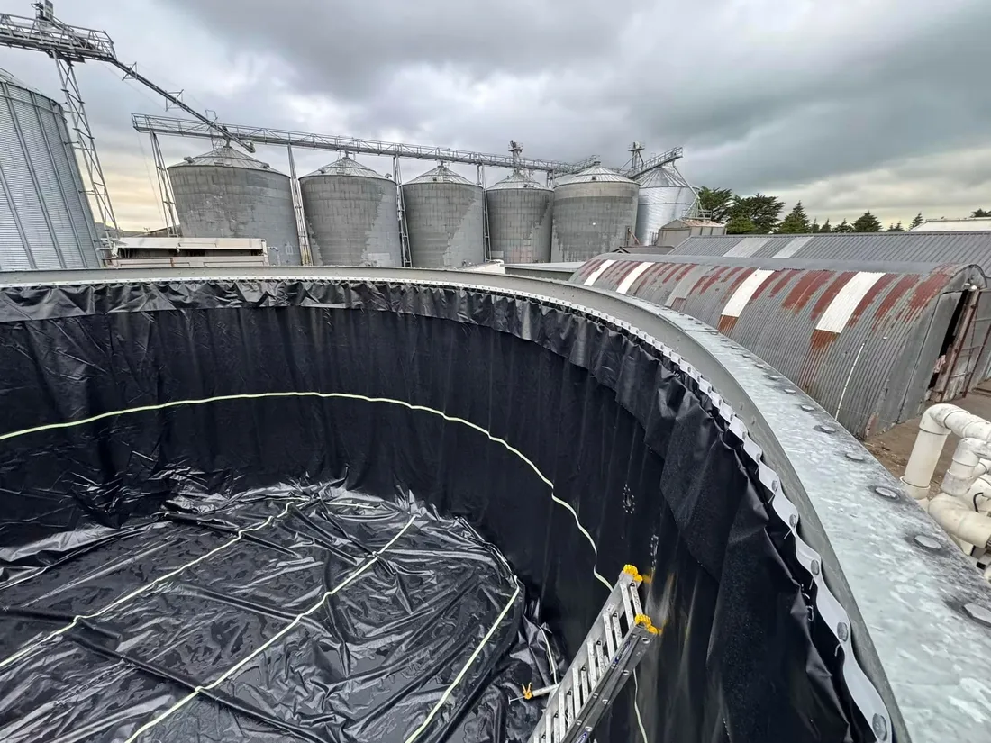 Black liner fitted inside a bolted steel tank on a rural site with grain silos behind