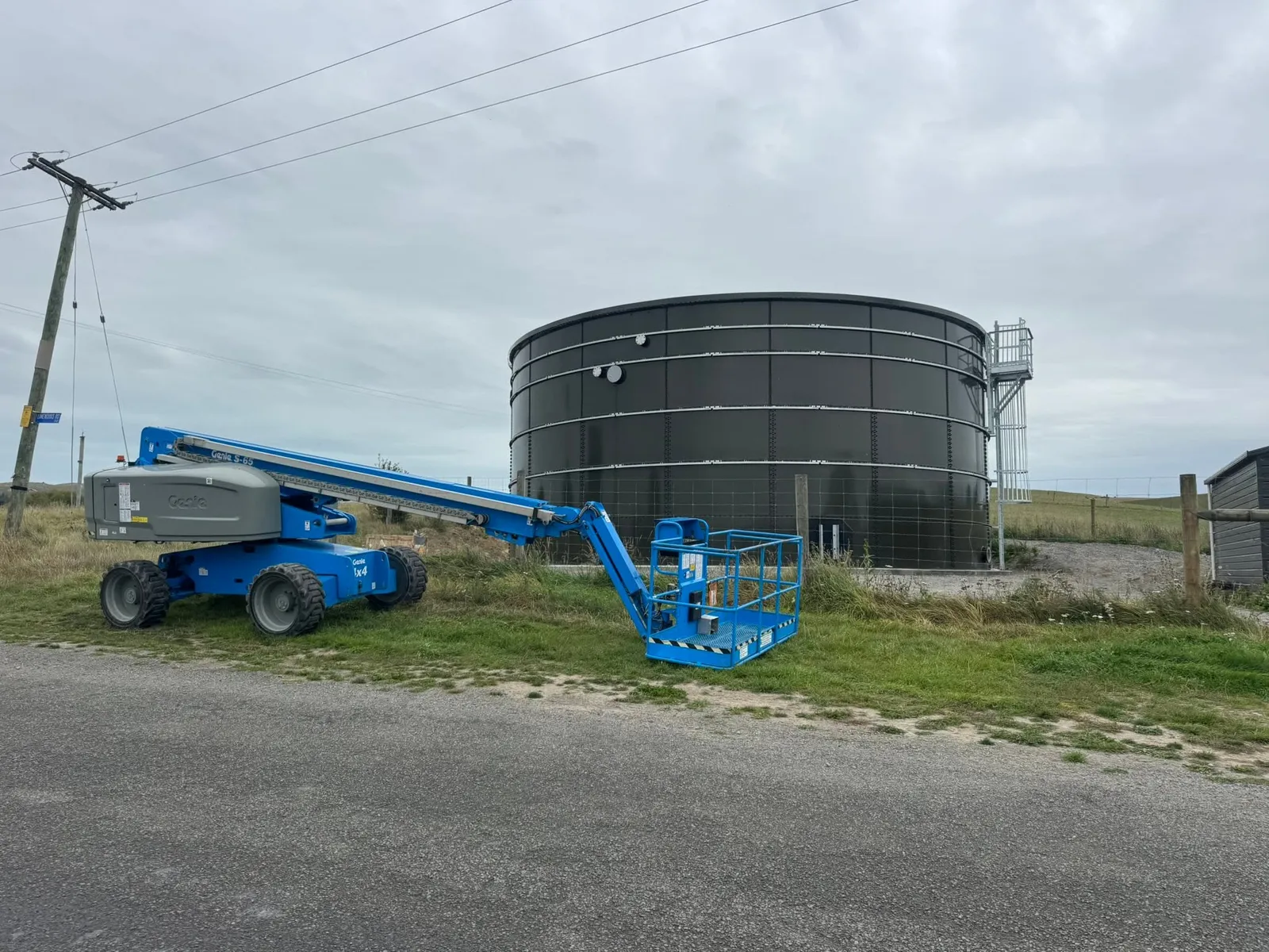 A boom lift set up beside a completed dark bolted steel tank on a rural site