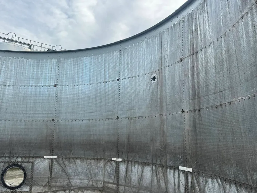 The inside wall of the tank stripped back to bare panels, water staining running down the plates, the rim open to an overcast sky