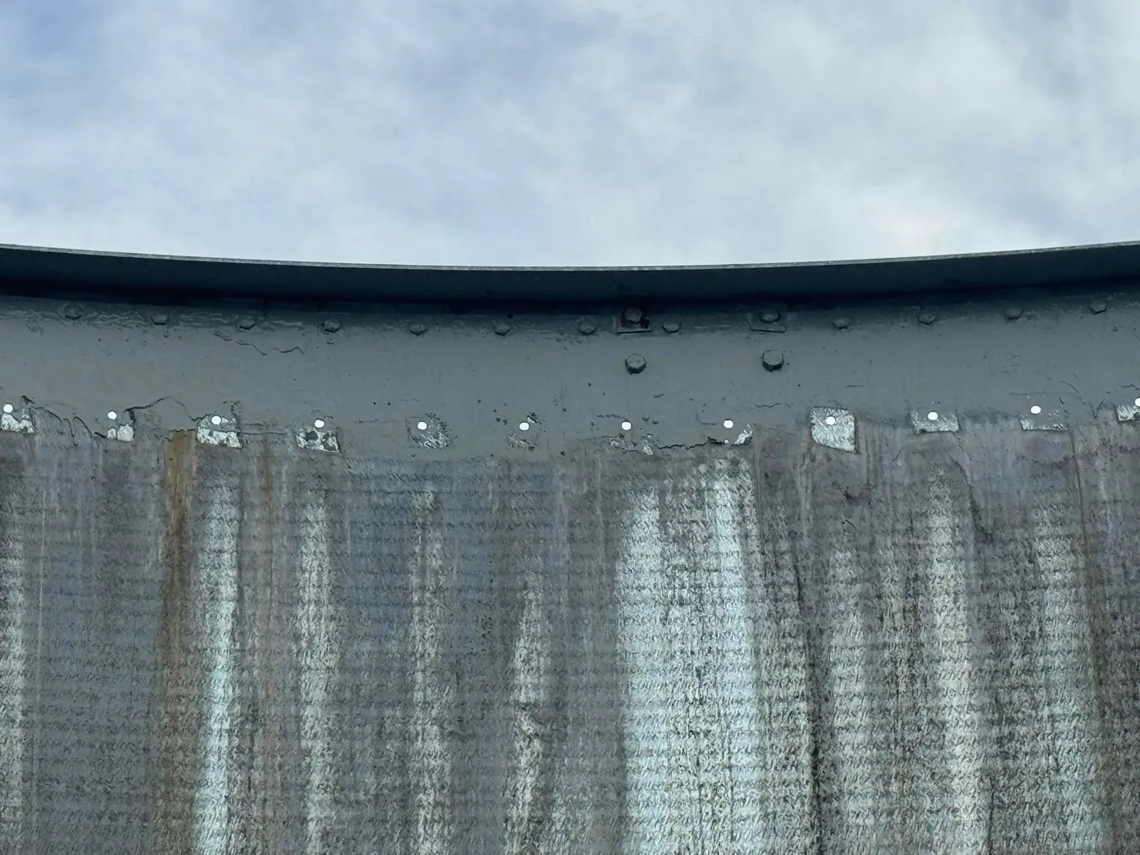 The weathered outside wall of the tank under an overcast sky, streaked the full height of the panels