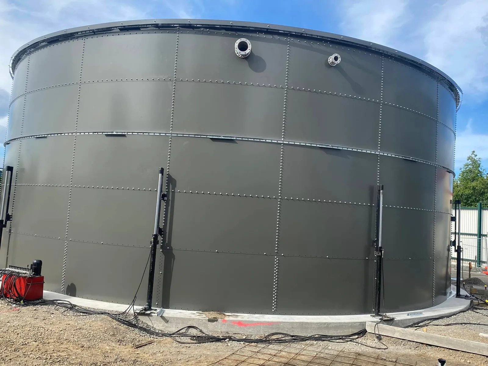 A bolted steel tank shell part built on its ring beam, hydraulic jacking rams standing against the wall